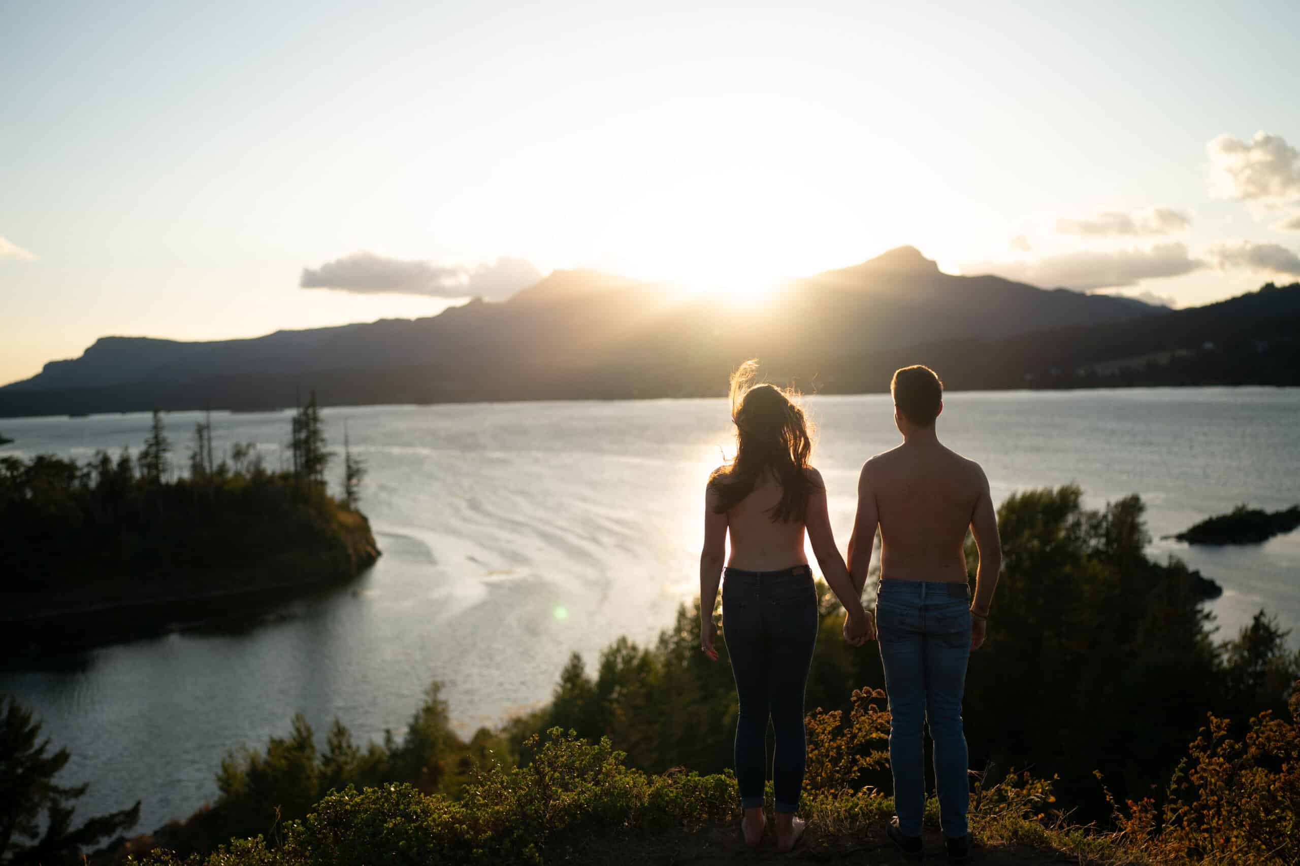 Couple holding hands, watching sunset over water.
