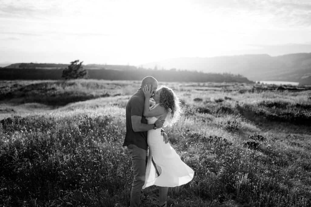 A couple embraces in a field at sunset. The woman, in a flowing dress, holds the man's face, while he wraps an arm around her waist. The background features rolling hills and an open sky, creating a romantic and serene atmosphere. Black and white photo. - Jessica Hill Photography
