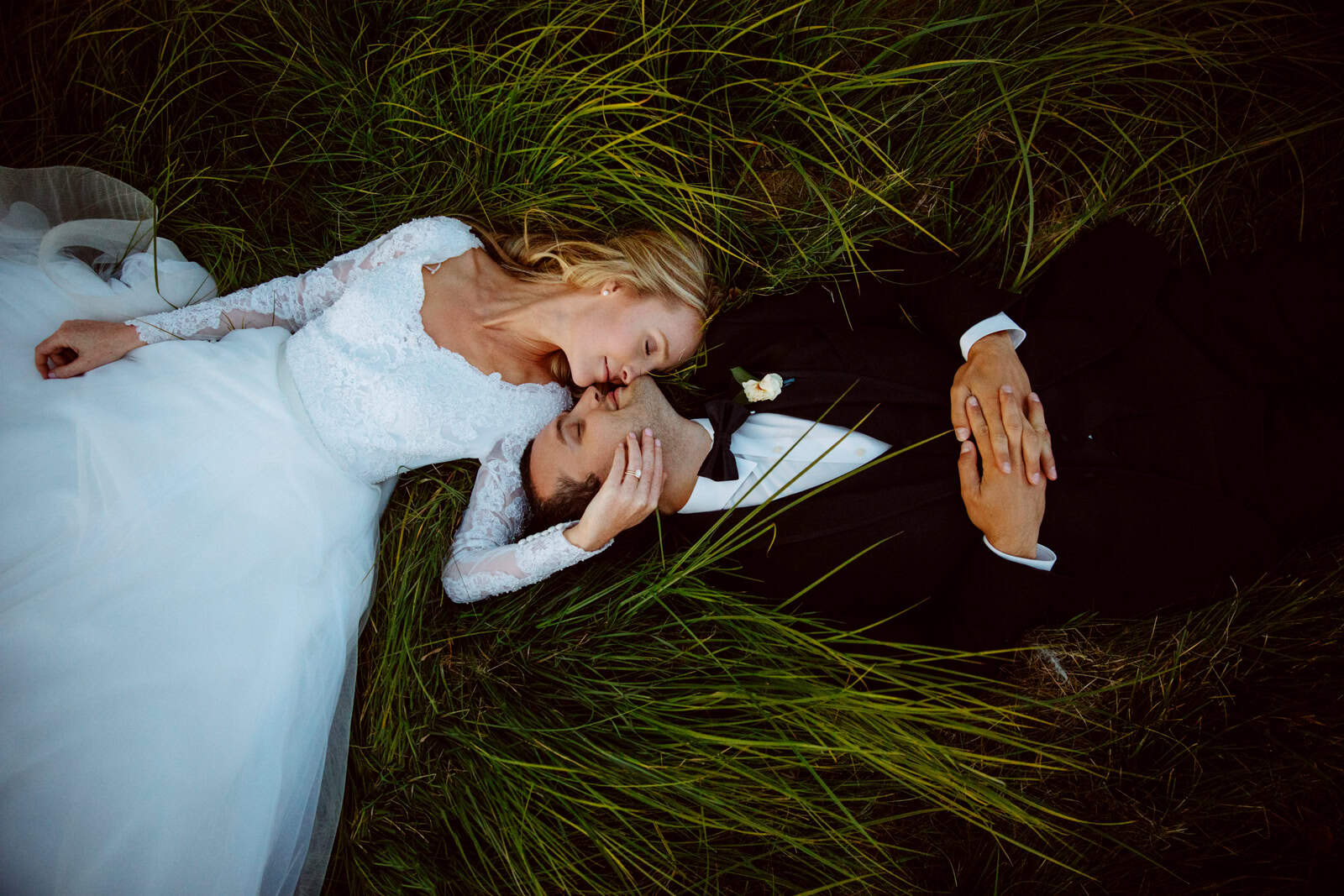 A bride in a white lace gown and a groom in a black suit lie on grass, heads close together. The bride is looking at the groom with a gentle smile while the groom has his eyes closed, creating a serene and intimate moment. - Jessica Hill Photography