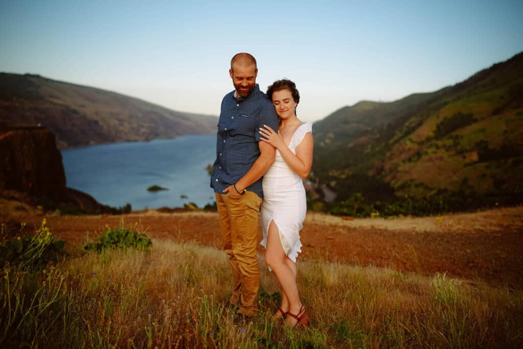 Couple standing on hill overlooking river and mountains.